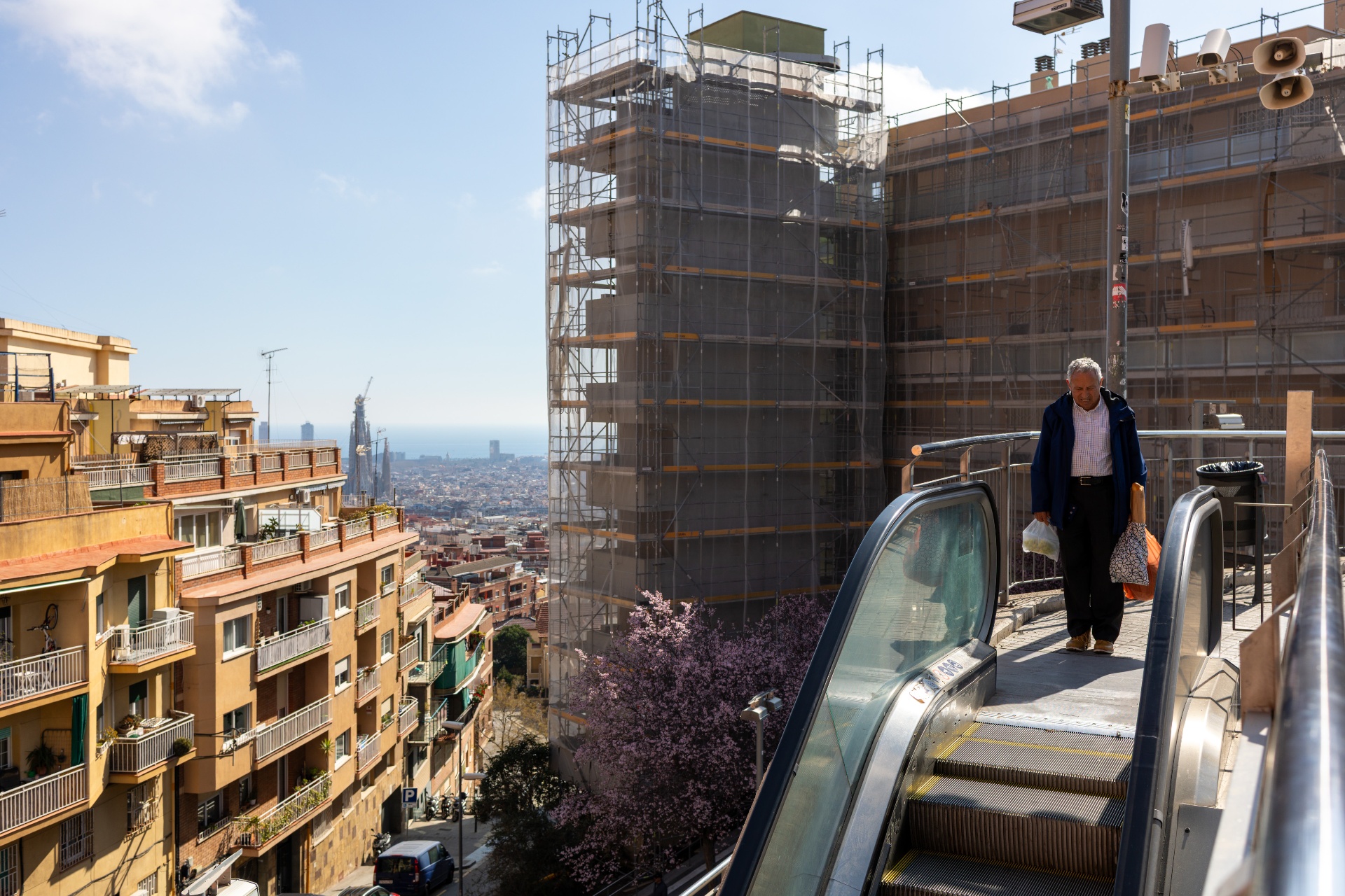 A man standing on an outdoor escalator above a sunlit Barcelona cityscape